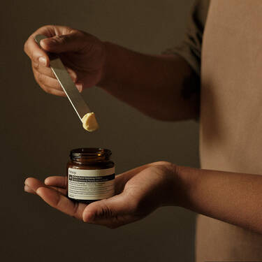An editorial, mid-shot photograph of a person holding an open amber glass jar of Aesop "Perfect Facial Hydrating Cream" in one palm. With their other hand, they use a sleek silver metal spatula to scoop out a small dollop of the thick, pale-yellow cream. The person is wearing a minimalist, earth-toned brown apron against a soft, dark-olive textured background.