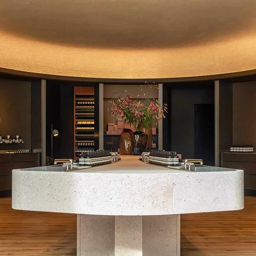 An elegant beauty boutique with minimalist design, featuring a large stone central island with integrated sinks and products, surrounded by dark wood shelving and warm ambient lighting.