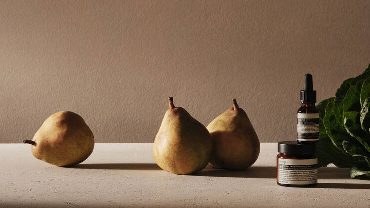 A minimalist still life featuring three pears, green leafy vegetables, and stacked Aesop skincare bottles on a textured beige surface against a warm, neutral background.