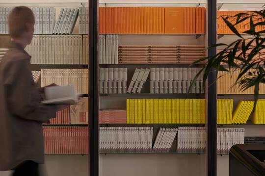 Blurred person walking past warm wooden shelves filled with yellow, orange, and red books.