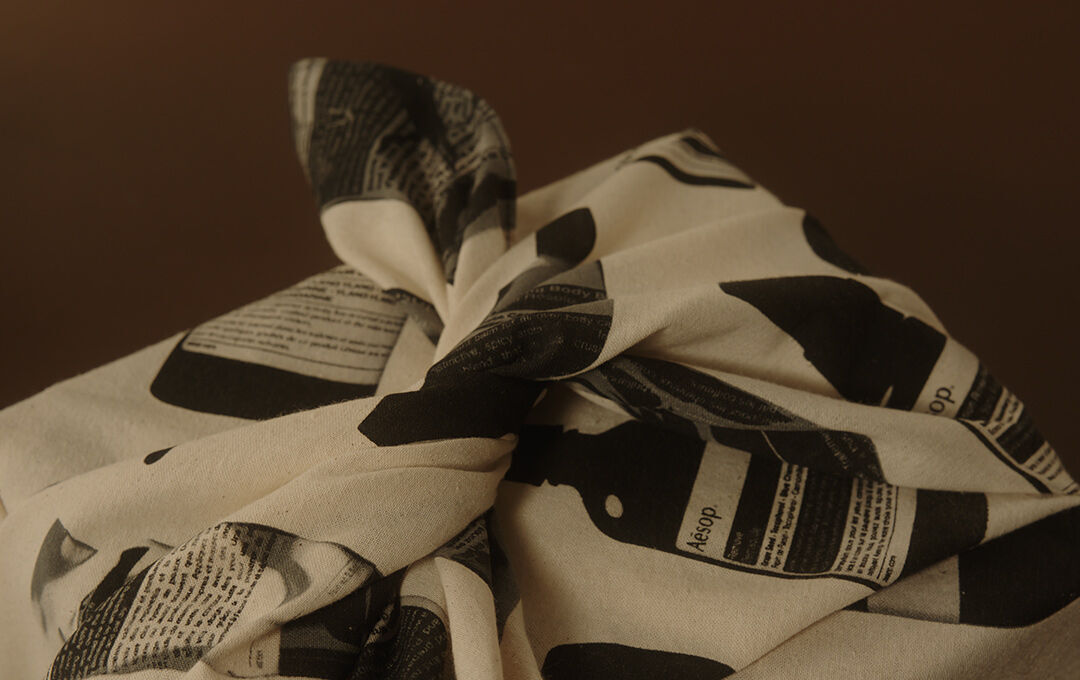 A close-up photograph of a package wrapped in a cream-colored cotton fabric using a traditional knotting technique (Furoshiki). The fabric is patterned with black-and-white graphics of Aesop bottles and text labels. The package is set against a warm, minimalist brown background under soft lighting.