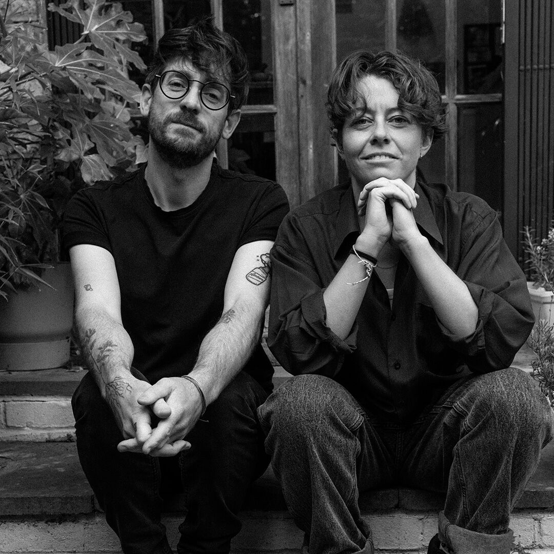 A black and white medium shot of two people sitting side-by-side outdoors. 