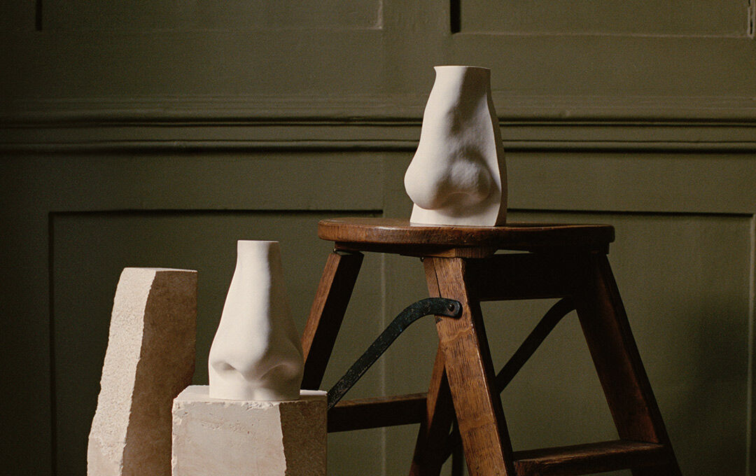 An artistic, still-life photograph featuring two white plaster sculptures of human noses. One sculpture rests on a rough, off-white stone block in the foreground, while the second is perched atop a rustic wooden step-stool. They are set against a muted olive-green, wood-panelled wall under soft, warm lighting.
