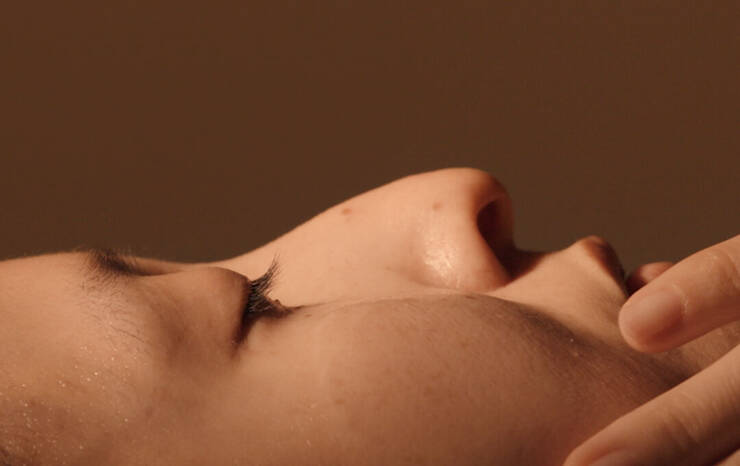 A close-up, side-profile view of a person lying down with their eyes closed. The warm, soft-focus lighting highlights the contours of their nose, long eyelashes, cheek, and upper lip. A hand is shown gently touching the lower part of their face near the chin, set against a solid, warm brown background.